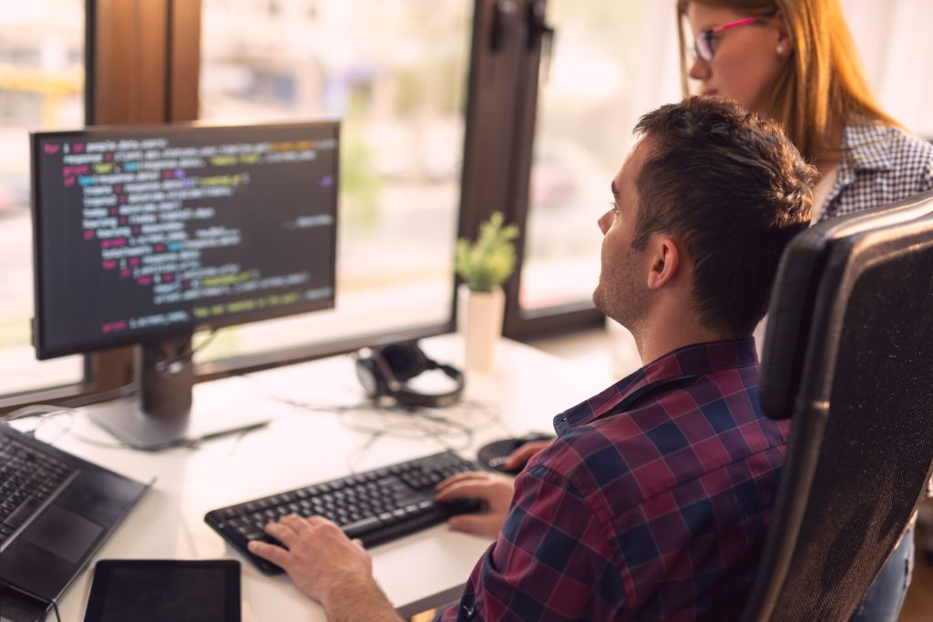 Dois desenvolvedores trabalhando em um escritório de uma empresa. Um homem sentado de frente para o computador, e uma mulher em pé. Ambos estão olhando para a tela, onde há bastante código.