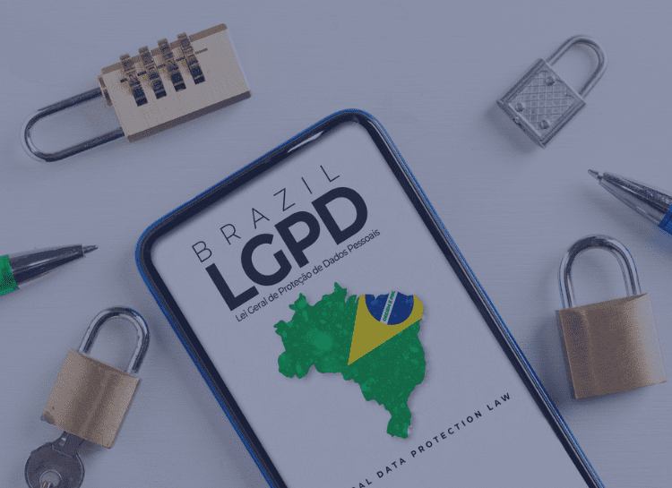 Na imagem vemos um celular sobre uma mesa, na tela está escrito Brazil LGPD, abaixo vemos um mapa com uma bandeira do país. Na mesa há alguns cadiados. A imagem foi escolhida para representar os princípios da LGPD.