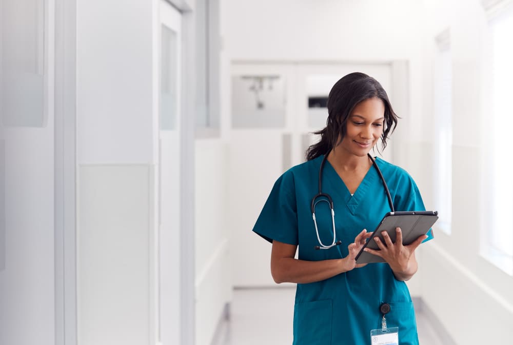 Médica no corredor do hospital segurando um tablet e sorrindo suavemente. Representando com esta imagem a importância dos hospitais se tornarem digitais para conquistar a certificação HIMSS.