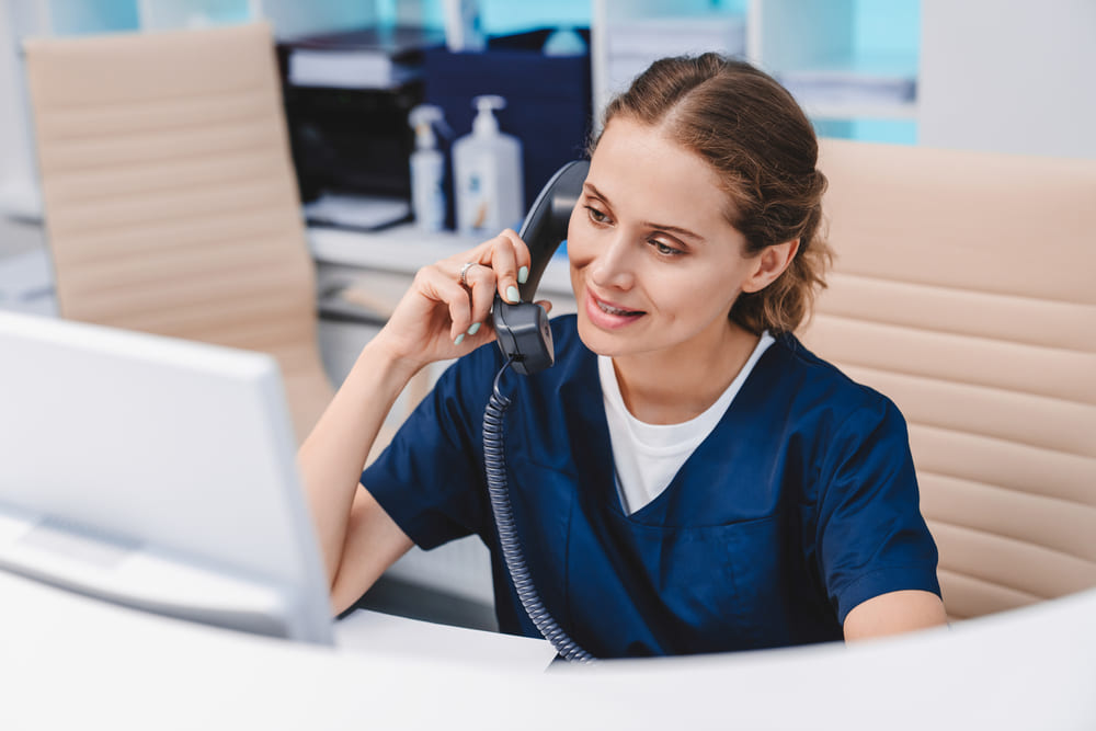 Enfermeira sentada atendendo o telefone e olhando para o computador. Ela usa um pijama cirúrgico azul e está sorrindo. Representa o profissional capaz de impulsionar a transformação digital na saúde.
